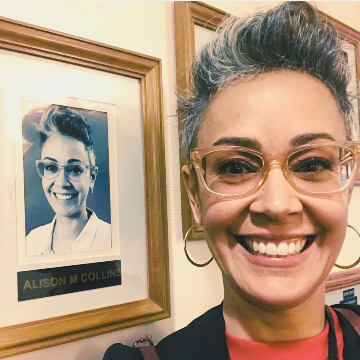 A headshot of Ali Collins in front of her black and white district profile photo hung on the wall. She is wearing peach colored glasses and smiling. Her hair is short and is wearing a red sweater, black blazer and large hoop earrings.